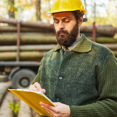 A man wearing a hard hat, standing in front of a truck with logs, reviewing a report.